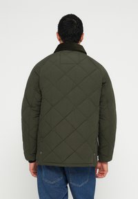 Casaco verde matelassado com gola de bombazina, ombros arredondados e corte descontraído, combinado com jeans azuis.