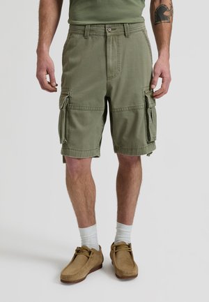 Olívazöld cargo rövidnadrágot, fehér zoknit és világosbarna, hétköznapi cipőt viselő férfi, karjait lazán a test mellett tartja, bal alkarján látható tetoválás.