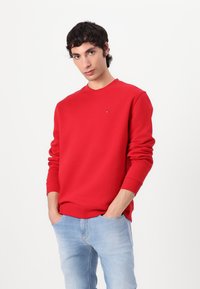 Rød crewneck sweatshirt i et blødt, tekstureret stof med ribbede manchetter. Har et lille logo på brystet. Sammen med lyseblå jeans.