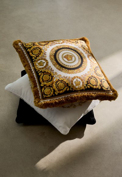 Square decorative pillows featuring a golden floral pattern on black, with a textured fringe. A white pillow is layered underneath.