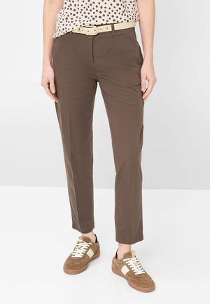 Person iført brune tapered bukser med beige bælte og brune ruskindssneakers med hvide accenter, stående på hvidt gulv.