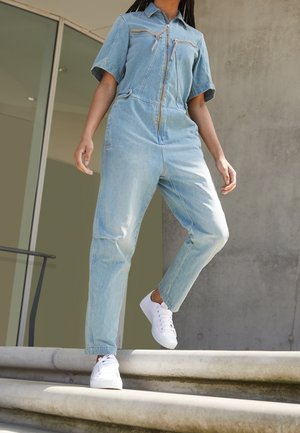 Jasnoniebieski kombinezon jeansowy z krótkimi rękawami, przednim zamkiem błyskawicznym i dwoma kieszeniami na piersi. Połączony z białymi trampkami na kamiennych schodach.