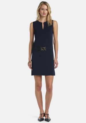 Szőke nő ujjatlan tengerészkék ruhában, elöl hasítékos nyakkivágással, derékban övvel, arany karika fülbevalóval, karkötővel és fekete T-pántos magas sarkúval.