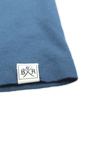 Blaues Baumwoll-T-Shirt mit abgerundetem Saum, versehen mit einem weißen Etikett mit schwarzer Schrift und gekreuzten Linien, die am unteren Rand eingenäht sind.