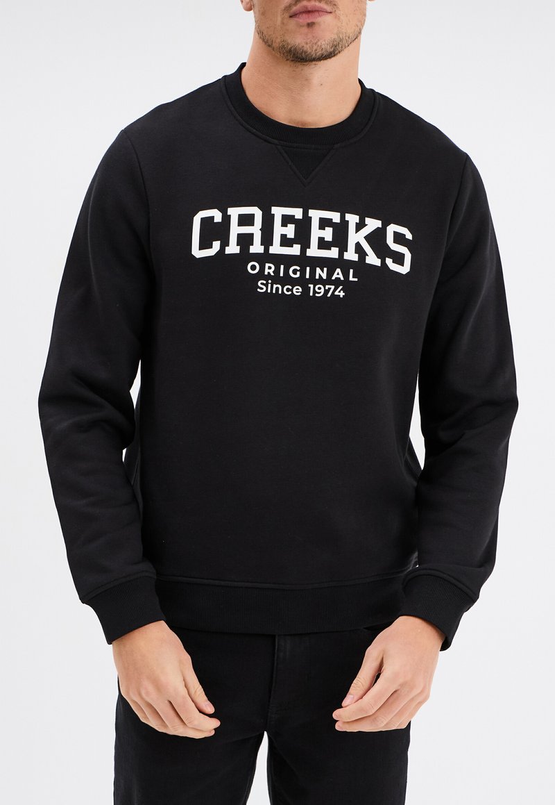 Sudadera negra con cuello redondo y puños de canalé. Presenta el texto "CREEKS ORIGINAL Desde 1974" impreso en blanco en la parte frontal.