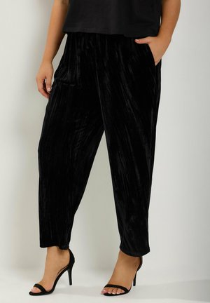 Persoon die losse zwarte fluwelen broek en zwarte hoge hakken met open teen draagt, staat met één hand in de zak tegen een eenvoudige achtergrond.