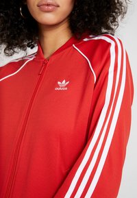 Červená zipová bunda vyrobená z hladké látky, s bílými pruhy na rukávech a malým bílým logem Adidas na hrudi.