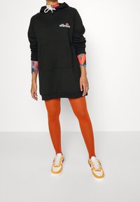 Svart huvtröjdress med framficka, kombinerad med orange leggings och vita sneakers med gula och lila accenter.