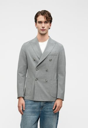 Un jeune homme se tenant debout, portant un blazer croisé gris clair sur une chemise blanche associé à un jean bleu, devant un fond uni.