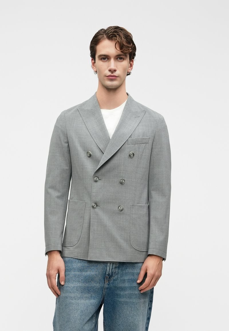 Νέος άνδρας που στέκεται φορώντας ένα ανοιχτό γκρι διπλό μπλέιζερ πάνω από άσπρη πουκάμισο, συνδυασμένο με μπλε τζιν, μπροστά σε απλό φόντο.