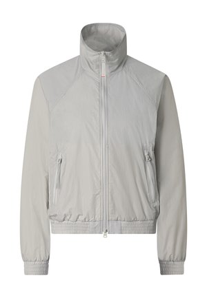 Frau trägt übergroße graue Reißverschlussjacke, passenden knielangen Rock, robuste Wanderschuhe und große reflektierende Visier-Sonnenbrille, steht aufrecht.