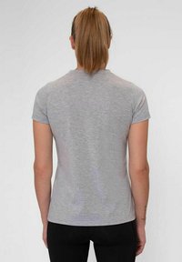 Vrouw met een bruine paardenstaart, gekleed in een effen grijze shirt met korte mouwen en zwarte broek, staand met haar gezicht naar een neutrale witte achtergrond.