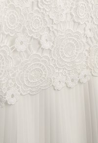 Weißes Kleid mit aufwendiger floraler Spitzenapplikation, die Pailletten enthält, darunter geschichteter plissierter Stoff und eine weiche, strukturierte Oberfläche.