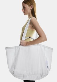 Liela balta iepirkumu soma, kas izgatavota no viegla auduma, ar pelēku apmalīti, garām siksnām un sānu logo etiķeti. Gluda tekstūra un plašs dizains.