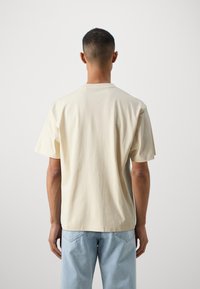 Tricou din bumbac bej deschis, cu mâneci scurte, gât rotund și o croială lejeră, văzut din spate. Fără modele sau grafica vizibile.