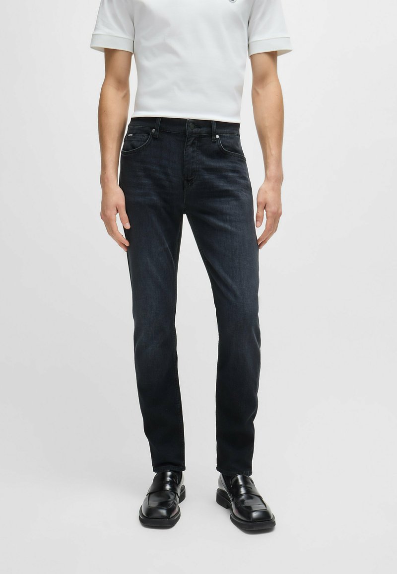 Dark blue denim jeans with a slim fit, featuring a standard five-pocket design and belt loops, paired with black loafers.