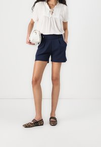 Femme portant une blouse blanche, un short bleu marine, des ballerines à imprimé léopard, tenant un sac à main blanc rond devant un fond uni.