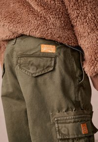 Gröna cargobyxor i en strukturerad väv, med en orange SuperDry-logotyp patch, fickor och en figursydd midja. Mysig brun fleeceöverdel som är delvis synlig.
