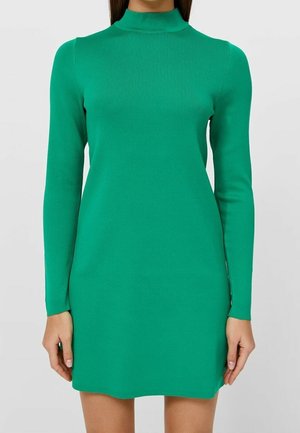 Kobieta w dopasowanej, długorękawowej, zielonej dzianinowej sukience z golfem, stoi na tle jednolitego jasnoszarego tła.