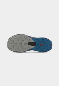 Suela de una zapatilla de trail running azul y gris con tacos multidireccionales y una "X" roja cerca del arco para tracción y estabilidad.
