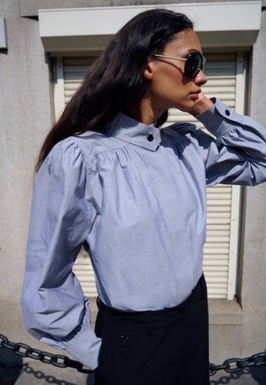 Chemise en coton bleu clair avec col montant, épaules plissées et détail de boutons. Manches longues avec poignets froncés. Pantalon noir visible.