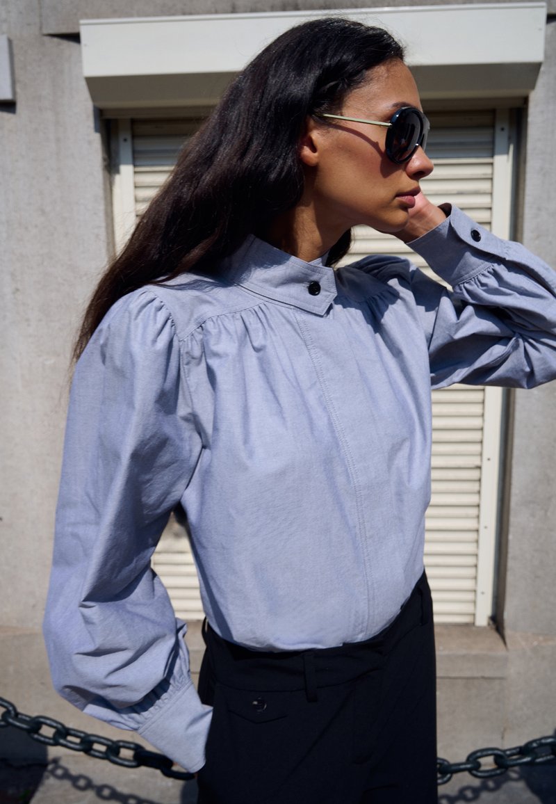 Lichtblauw katoenen shirt met een hoge kraag, geplooide schouders en knoopdetail. Lange mouwen met gerimpelde manchetten. Zwarte broek zichtbaar.