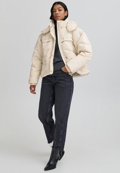 Sieviete valkā bēša krāsas polsterētu jaku pār tumšu džemperi, melnas augstās jostas džinsas un melnus smailus potīšu zābakus, stāv vienai rokai kabatā.