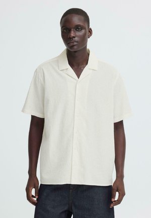 Homme portant une chemise à manches courtes, boutonnée, texturée et de couleur ivoire avec col ouvert, ainsi qu'un pantalon bleu foncé, debout devant un fond uni.