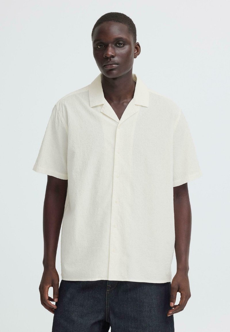 Homme portant une chemise à manches courtes, boutonnée, texturée et de couleur ivoire avec col ouvert, ainsi qu'un pantalon bleu foncé, debout devant un fond uni.