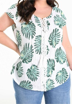 Woman wearing white short-sleeve blouse with green tropical leaf print and lace-up front detail, paired with blue jeans.