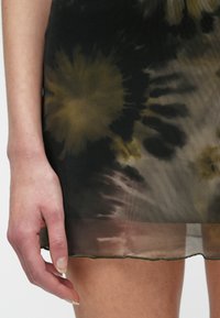 Leva roka počiva ob stegnu, oblečena v krilo iz prozorne tkanine v črno-rumeni tie-dye vzorcu z zavitim robom.