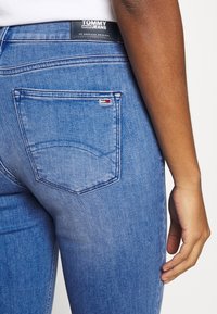 Närbild av en person som bär blå Tommy Jeans denim, som visar bakfickan med liten flagglogotyp och varumärkets etikett på midjebandet.