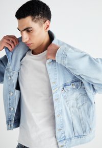 Chaqueta de mezclilla azul clara con cierres de botones, bolsillo en el pecho y ligero efecto de desgaste. Lleva puesta una camiseta blanca sencilla.
