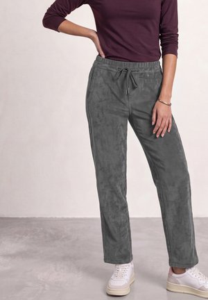 Personne portant un pantalon en velours côtelé gris avec cordon, des baskets blanches et un haut violet à manches longues, debout avec une main sur la hanche.
