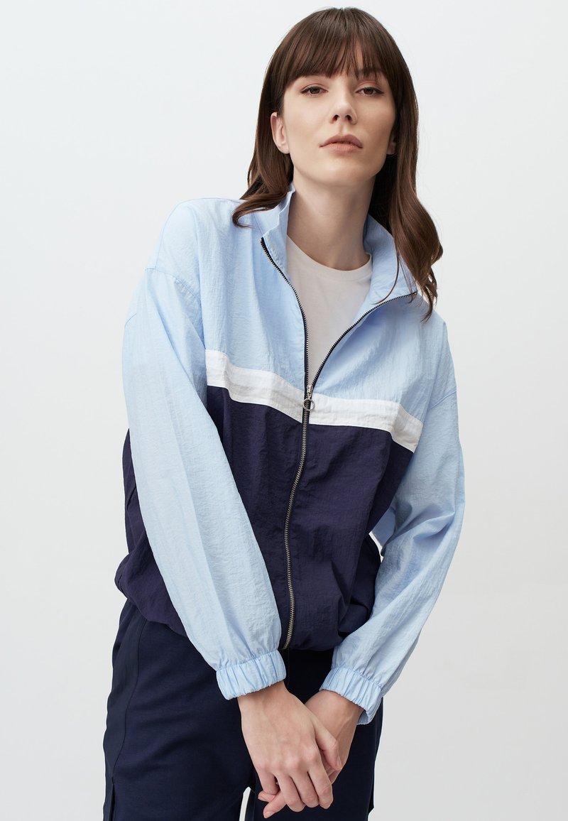 Giacca zip-up leggera blu chiaro e blu navy con una striscia orizzontale bianca. Presenta polsini elasticizzati e una vestibilità ampia, realizzata in un materiale leggero.