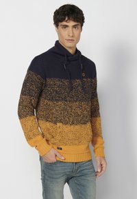 Jersey de punto con un degradado en azul marino y dorado. Cuenta con un cuello alto, cordón y un bolsillo frontal. Puños y dobladillo acanalados.