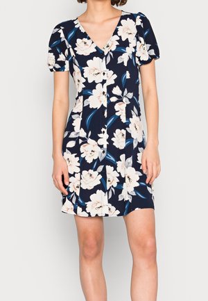 Žena oblečená v krátkých rukávech tmavě modrých šatech s bílým květinovým vzorem, výstřihem do V a knoflíky na přední straně.