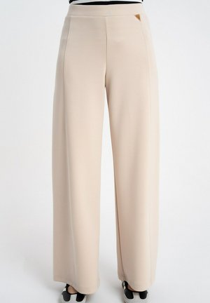 Beżowe spodnie o szerokich nogawkach z przeszyciami z przodu, noszone z czarno-białymi sneakersami, widoczne od talii do stóp.