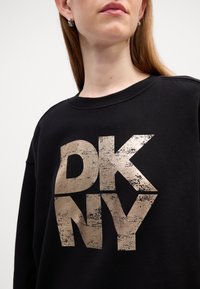Svart hettegenser med et stort, slitt gull"DKNY"-logo. Klassisk rund hals-design med ribbede mansjetter og myk stofftekstur.