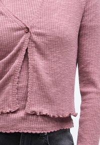 Ροζ μακρυμάνικο ριμπ τοπ με λαιμόκοψη σε σχήμα V, με ένα κουμπί κλεισίματος, υφή υφάσματος και λεπτομέρειες με κυματιστό τελείωμα.