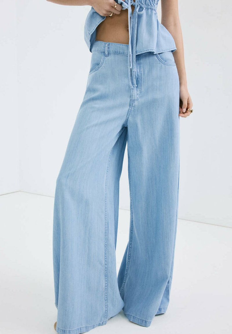 Vrouw draagt lichtblauwe wijdvallende jeans en een bijpassende denim top met koord, staand tegen een effen witte achtergrond.