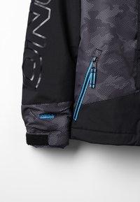 Svart jacka med texturerad tyg, prickigt mönster, blå dragkedjedetaljer och logotypdetaljering på ärm och bröst. Har justerbara ärmslut.