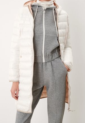 Stojící osoba nosící dlouhý bílý péřový kabát přes šedou texturovanou mikinu na zip a odpovídající kalhoty, s jednou rukou v kapse.