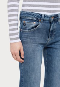 Jeans din denim într-o nuanță medie, având un design clasic cu cinci buzunare, cu nituri de cupru și un talie texturată, cu o mână odihnindu-se pe latură.