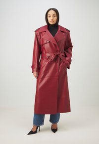 Rød faux læder trench coat med dobbeltknaplukning, bred krave, bælte i taljen og skulderepaulettes. Lagdelt over blå jeans med opsmøg.