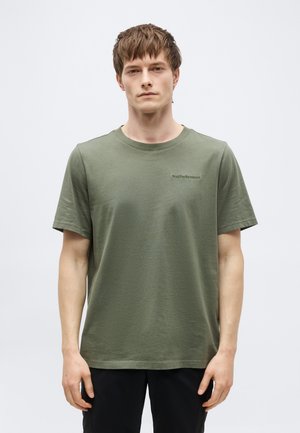 Giovane uomo che indossa una t-shirt a maniche corte verde oliva con un logo sottile sul petto, in piedi davanti a uno sfondo chiaro e uniforme.