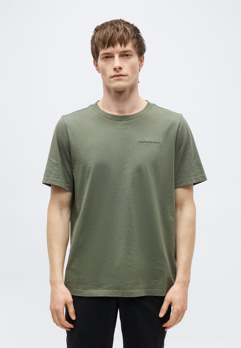 Jeune homme portant un t-shirt à manches courtes olive avec un logo subtil sur la poitrine, debout devant un fond clair uni.