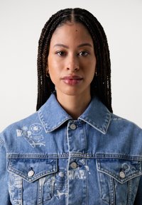 Veste en denim bleu clair avec des graphismes blancs imprimés sur le devant. Elle présente un col classique et des fermetures à boutons-pression.