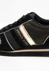 Sneaker Versace Jeans noire avec accents dorés, détails en mesh et lacets noirs sur une semelle en caoutchouc noire.