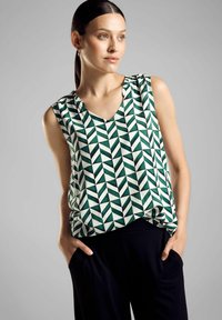 Top senza maniche con scollatura a V in un motivo geometrico verde e bianco, realizzato in un tessuto morbido, abbinato a pantaloni scuri e larghi.